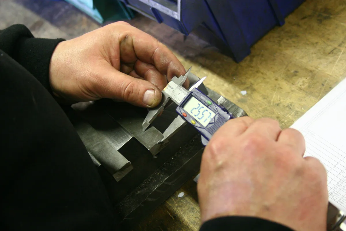 Hands measuring a machined metal part with a digital caliper on a workbench
