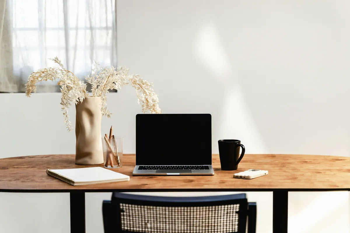 A minimal wooden desk with a laptop, a mug, and a vase of dried flowers
