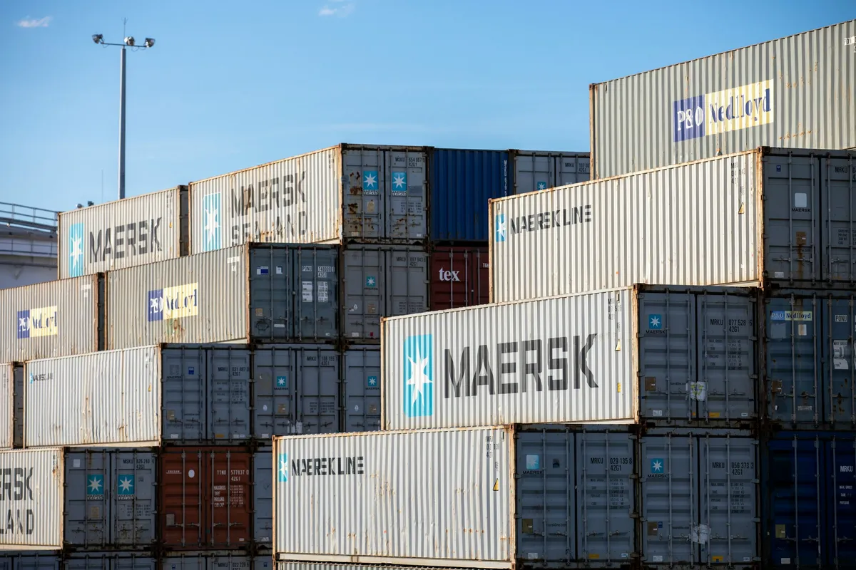 Stacks of grey and white shipping containers under a clear blue sky at a freight terminal