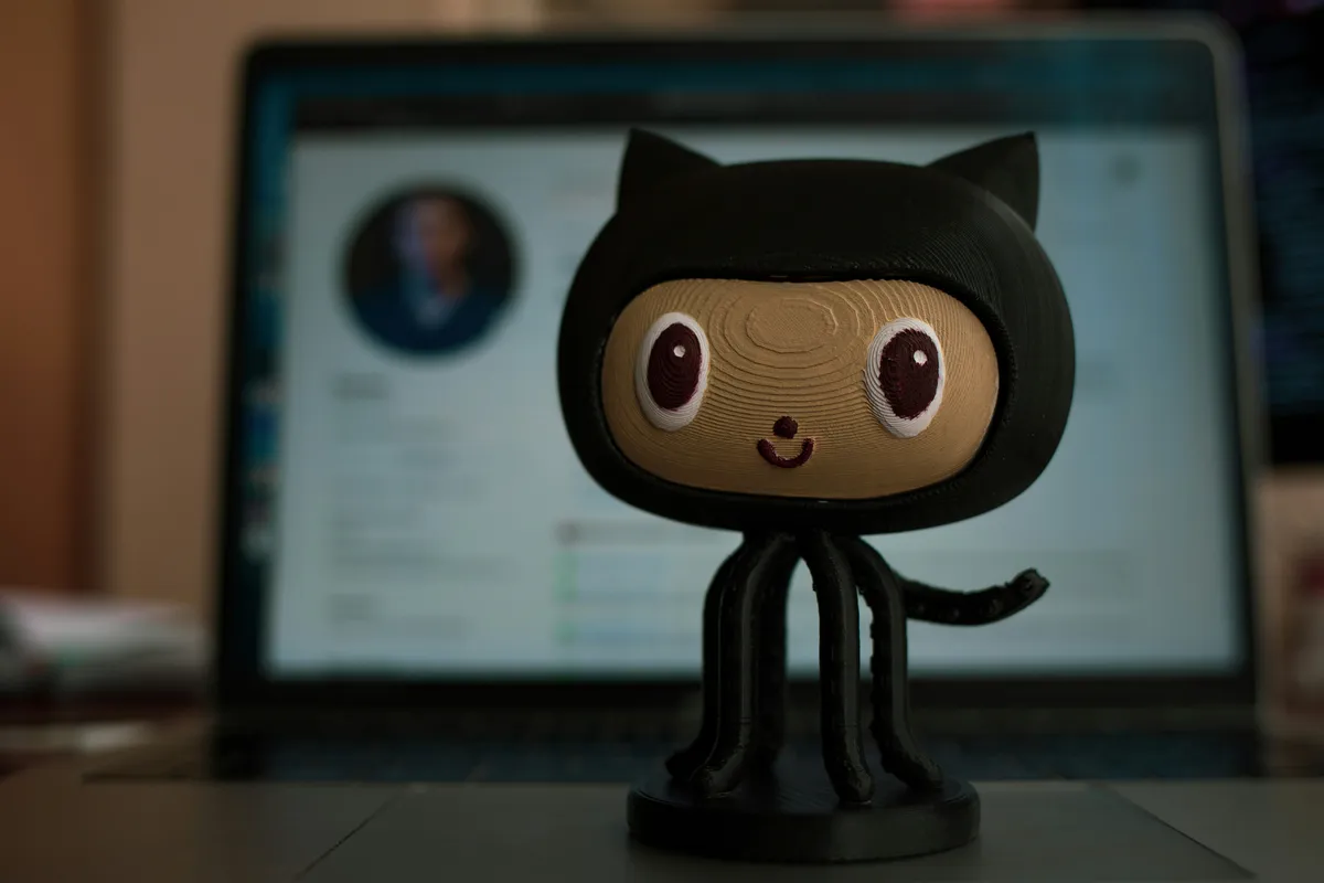 Octocat figurine standing in front of a laptop showing a blurred GitHub profile page