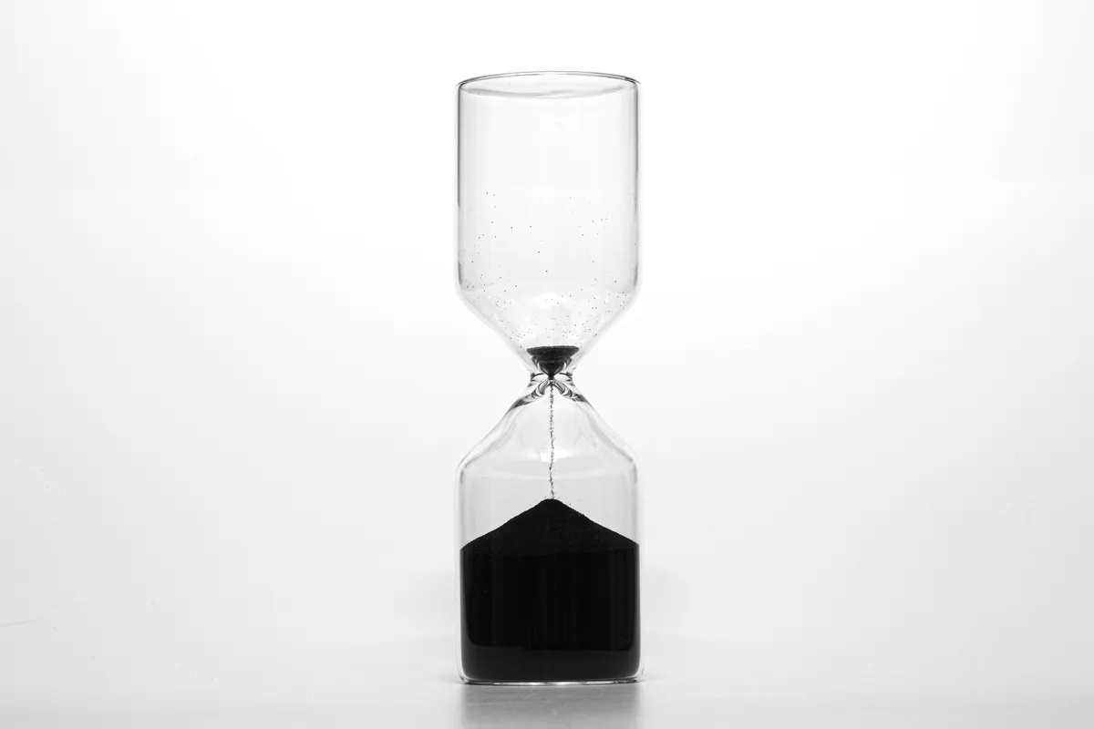 A glass hourglass with black sand running into the lower chamber on white